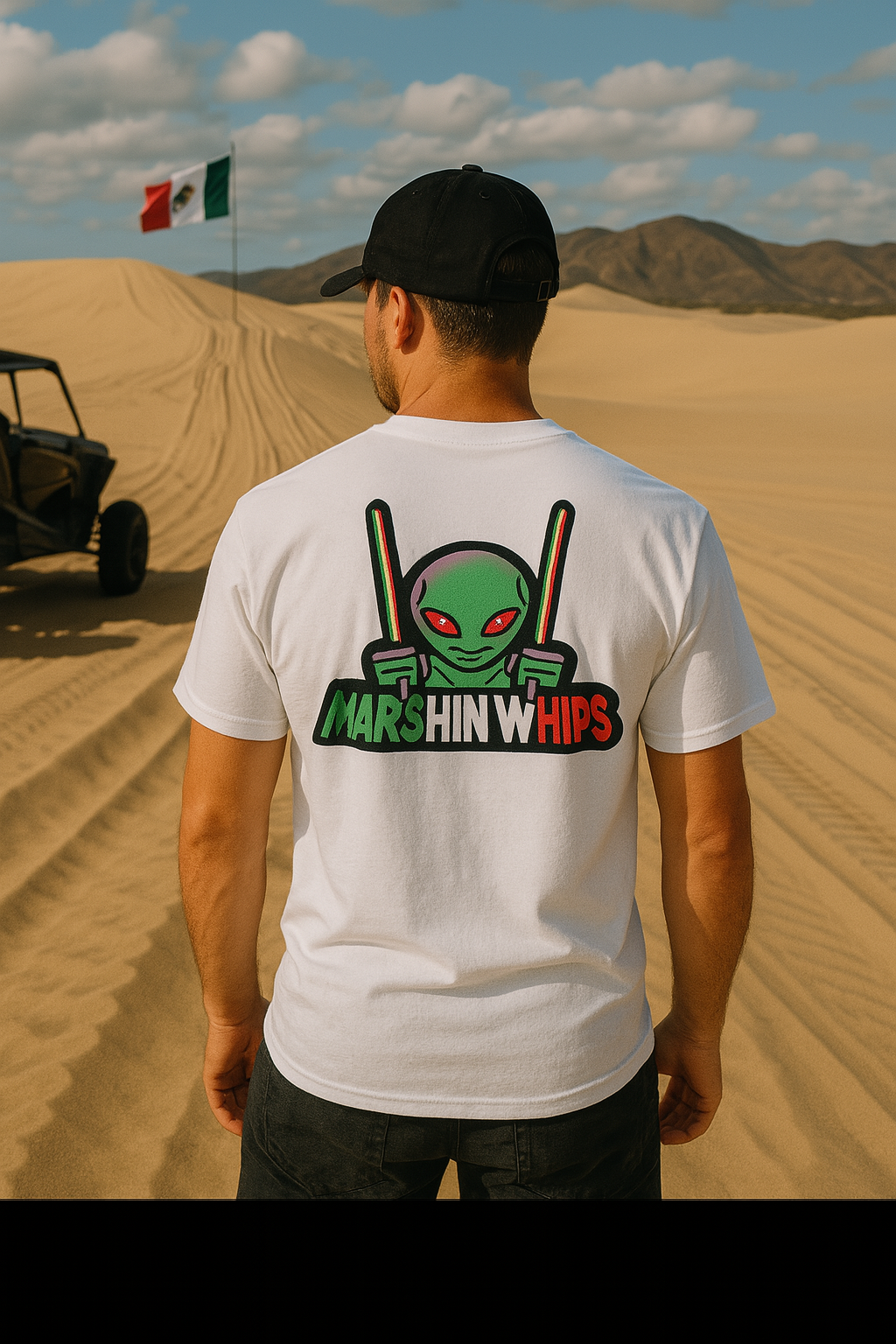 Back view of a man wearing a white "Marshin Whips" t-shirt with a large graphic of a green alien holding red, white, and green whip lights. He stands in the Mexican desert near an ATV with a waving Mexican flag and sand dunes in the background under a partly cloudy sky.
