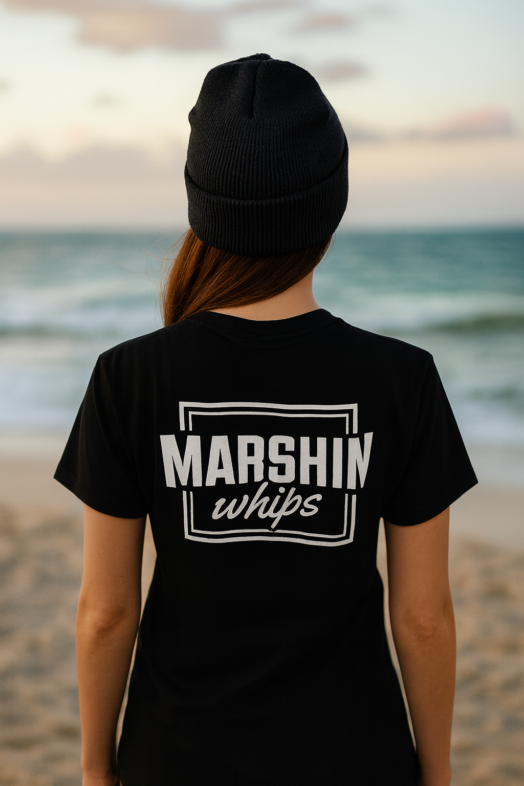 Person wearing a black t-shirt and beanie with 'MARSHIN whips' logo on a beach