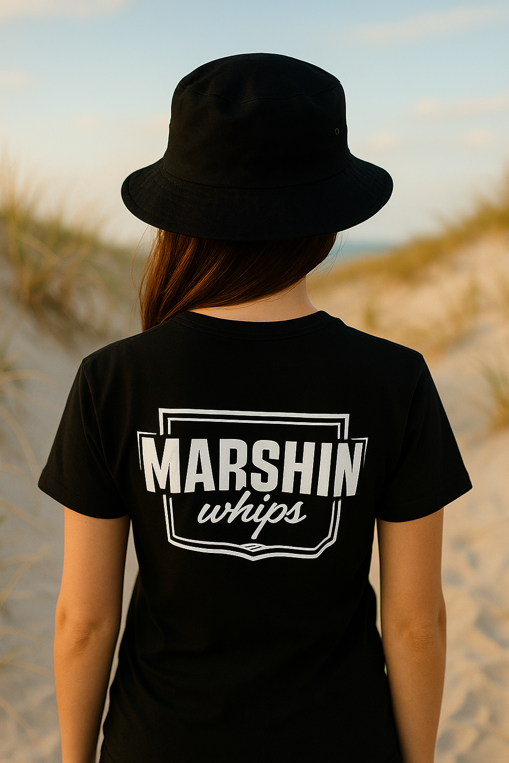 Person wearing a black t-shirt with 'MARSHIN whips' logo with a black bucket hat on a beach