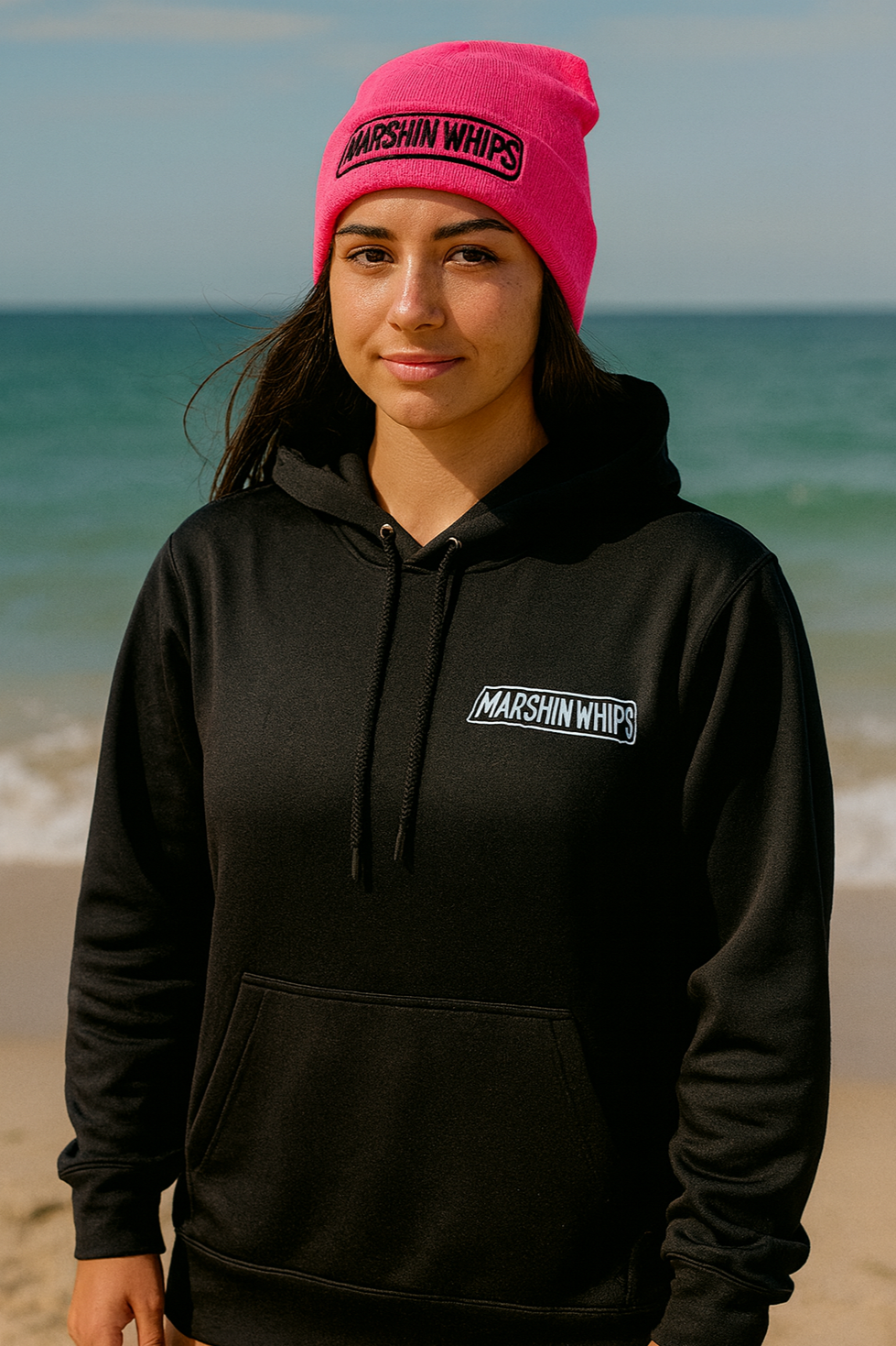 Person wearing a black hoodie and pink beanie with 'Marshin Whips' branding on a beach.