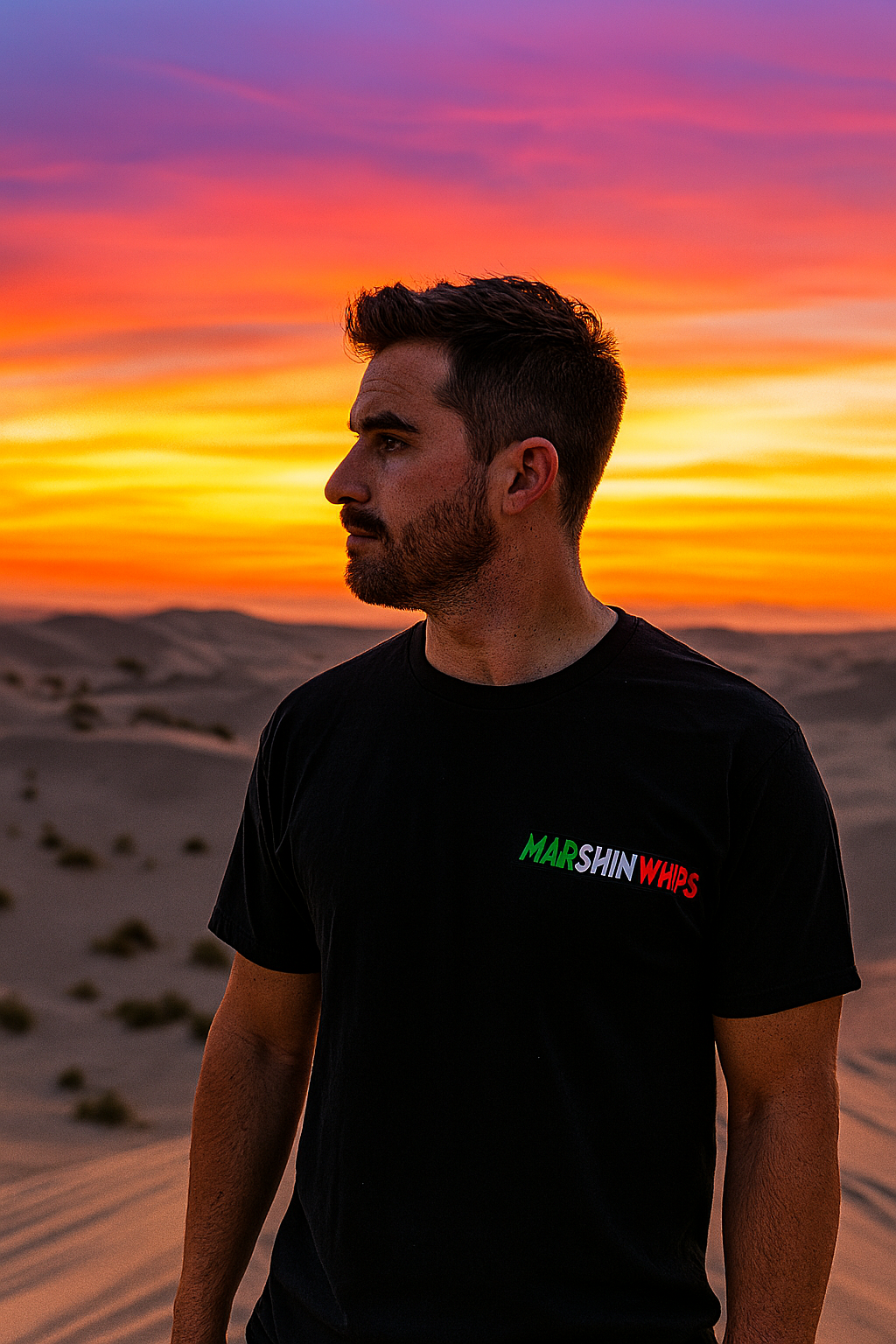 Man wearing a black "Marshin Whips" t-shirt with a small front logo in green, white, and red gradient text for Mexican Pride. He stands in the desert at sunset with a vivid orange, yellow, and pink sky glowing behind him.