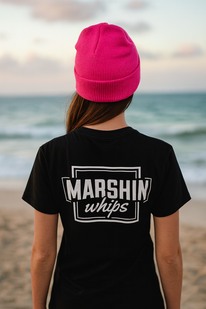 Person wearing a black t-shirt with 'MARSHIN whips' logo and a pink beanie on a beach.